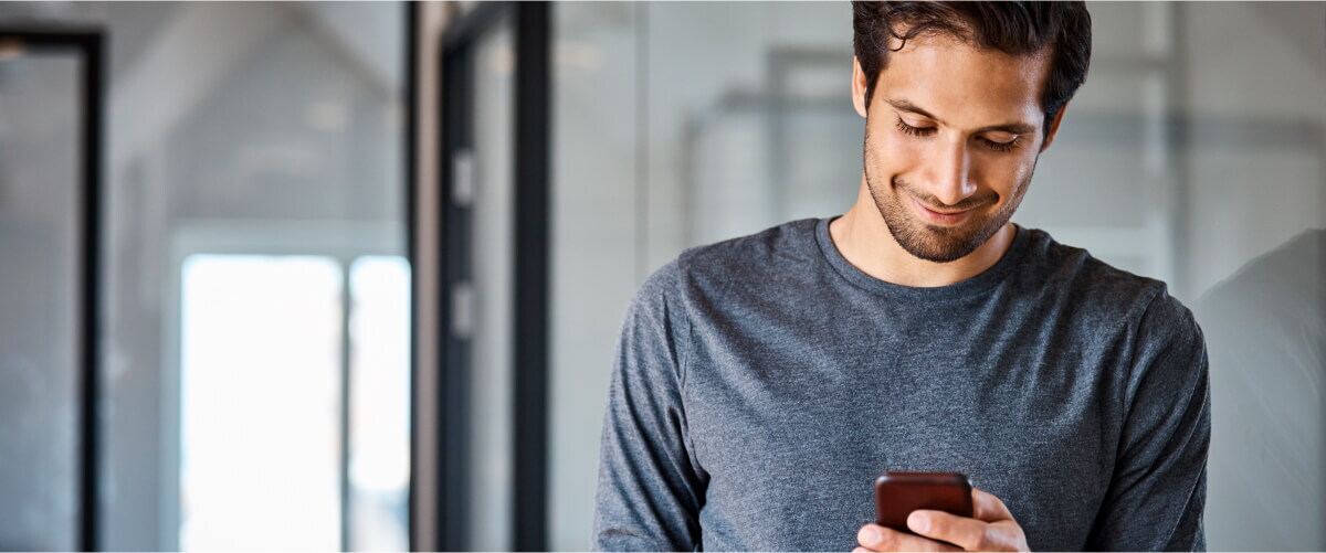 A smiling man in a grey shirt looks down at his smartphone while standing in a modern office