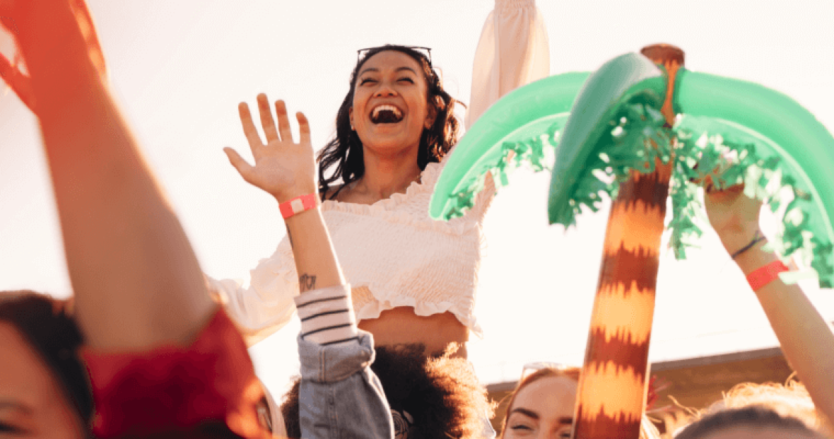 Person lifted above crowd at outdoor event, smiling with hands raised near inflatable palm tree.