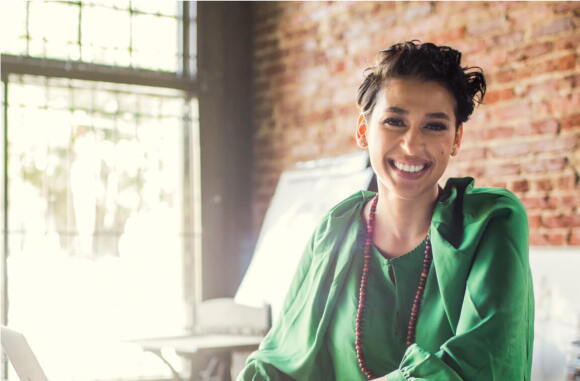 A smiling woman sitting inside a sun-lit office.