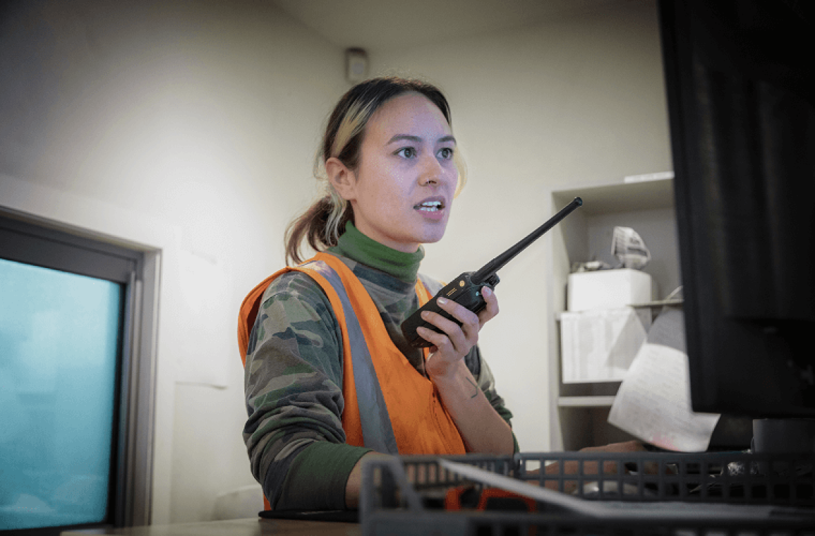Person in safety vest using walkie-talkie near computer workstation.