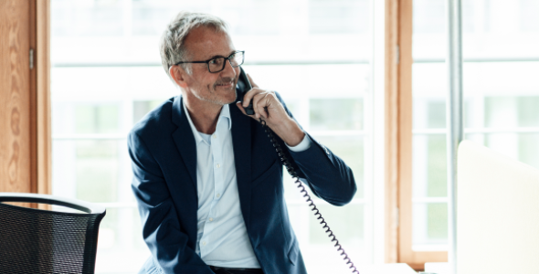 A smiling businessman wearing glasses and a suit talks on a corded office phone while seated beside a desk in a bright office.