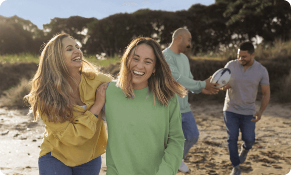 Two women laughing together on a beach while two men walk behind them, one holding a rugby ball.