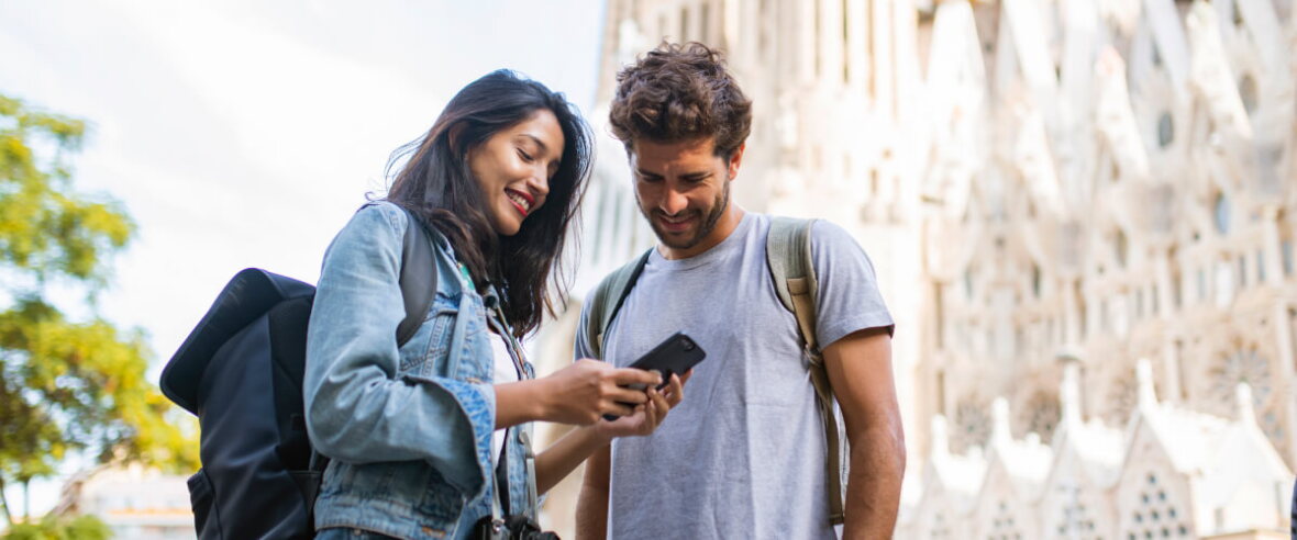 Two travellers with backpacks smile as they look at a smartphone together outside a large historic building.