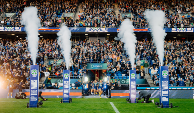 Rugby league players (One NZ Warriors) run onto the field through smoke effects in a packed stadium, with fans cheering and One NZ branding visible.