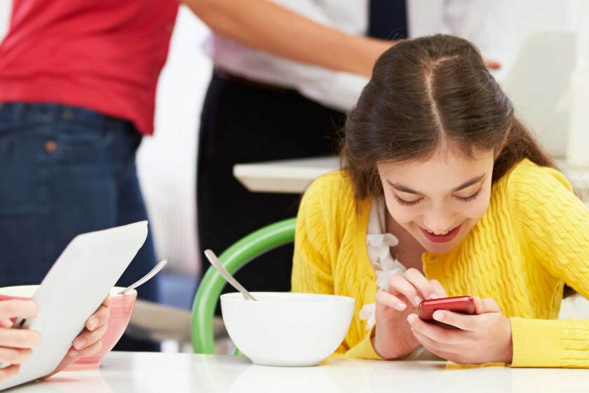 Two kids on devices at the breakfast table.