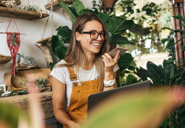 A smiling lady using her phone and laptop in her shop.