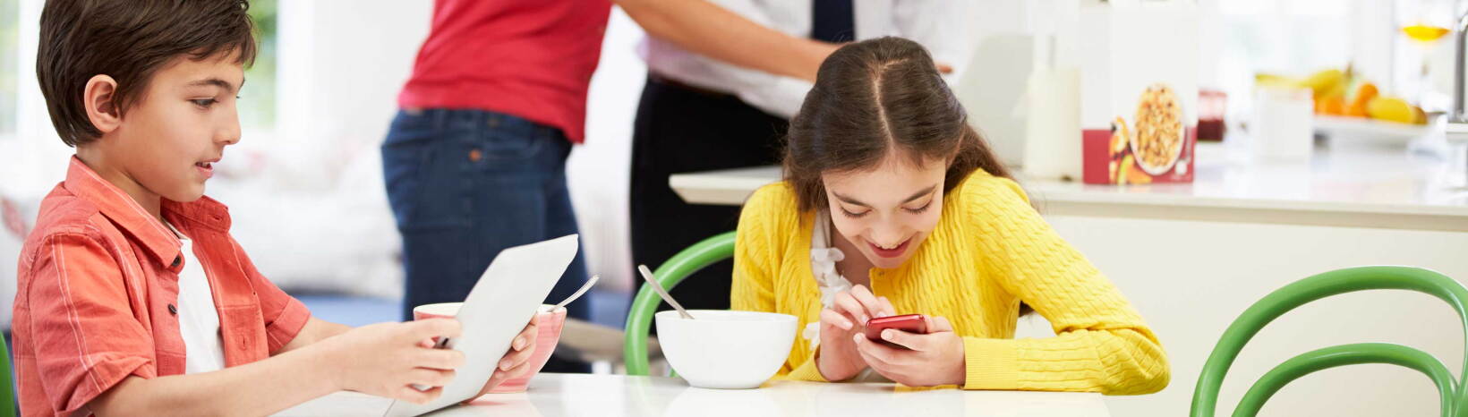 Two kids on devices at the breakfast table.