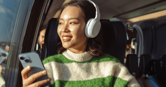 A young smiling woman looking out of a bus window, while holding her iPhone.