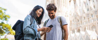 Two travellers with backpacks smile as they look at a smartphone together outside a large historic building.