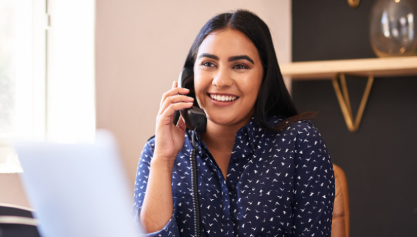 A smiling woman talks on a corded phone while seated at a desk in a modern office.