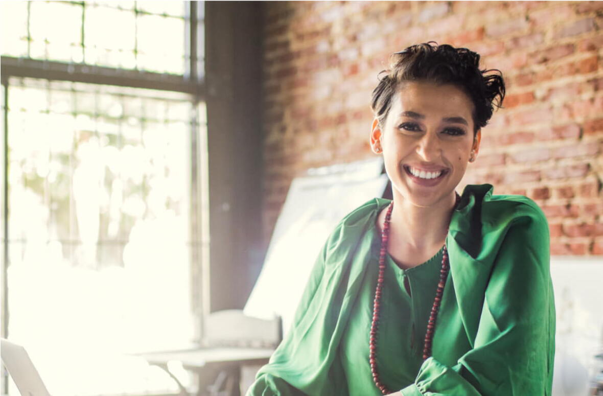 A smiling woman sitting inside a sun-lit office.