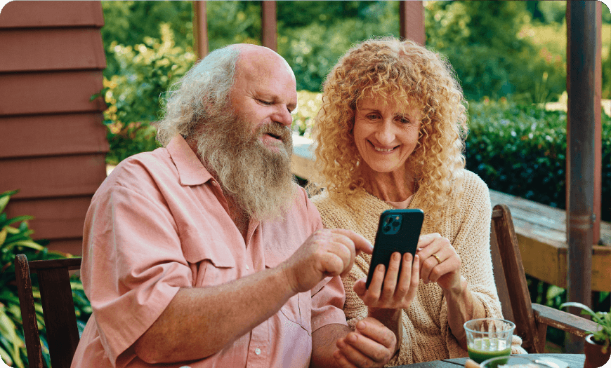 Older couple sitting outdoors and smiling while looking at a smartphone together.