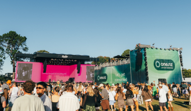 Large outdoor music festival crowd gathered in front of a stage with bright screens and One NZ branding.