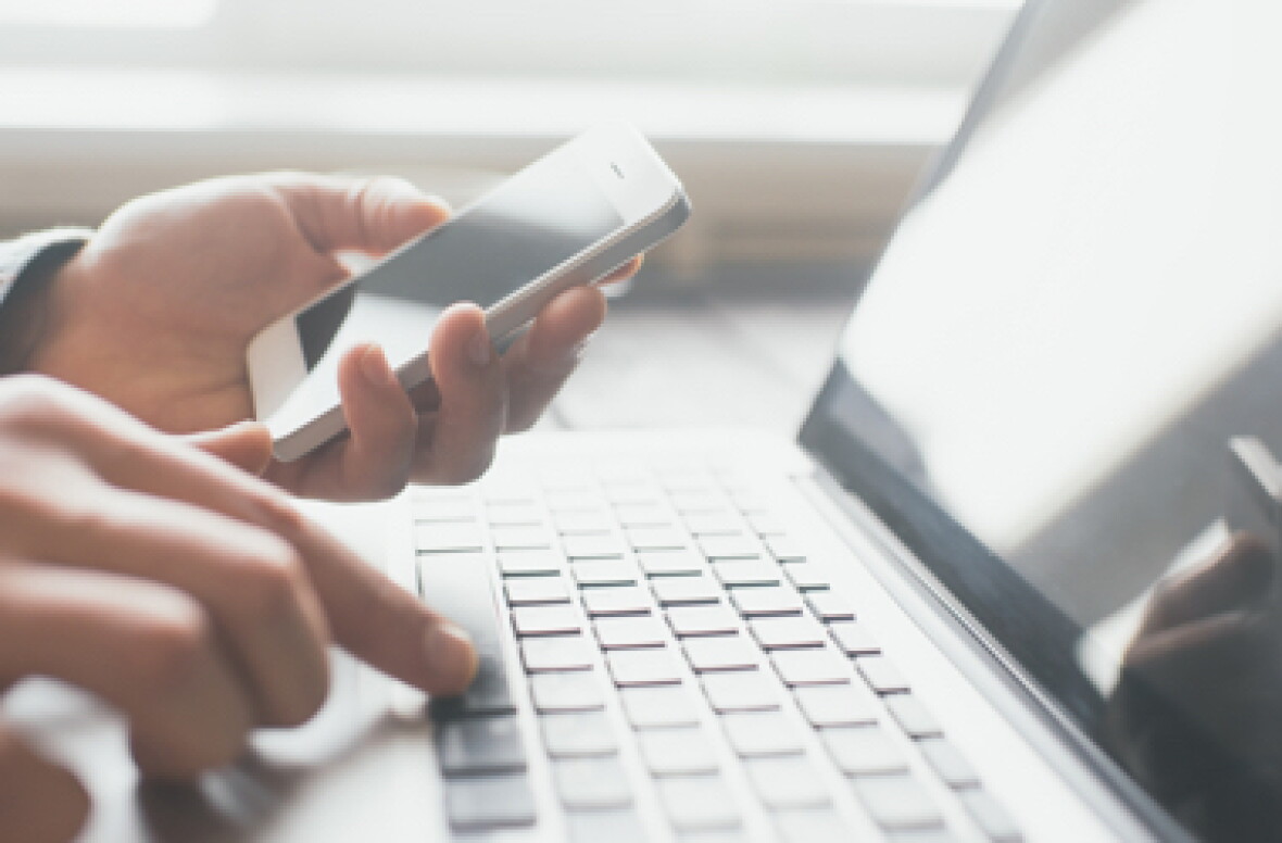 A person typing on a laptop while looking at a mobile phone