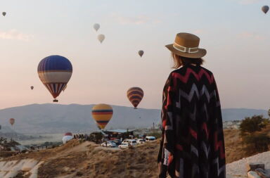 A lady wearing a wide-brimmed hat and patterned poncho watches colorful hot air balloons floating over a rocky valley at sunrise.