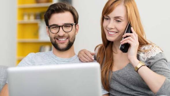 Two people sitting together using a laptop. One of them talking on phone.