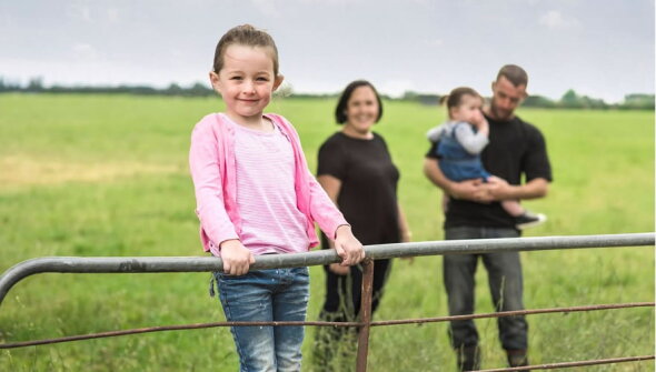 Girl wearing pink jersey standing on a farm gate with family behind her smiling.