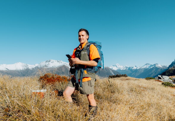 Person in grassy field with snowy mountains and clear blue sky.