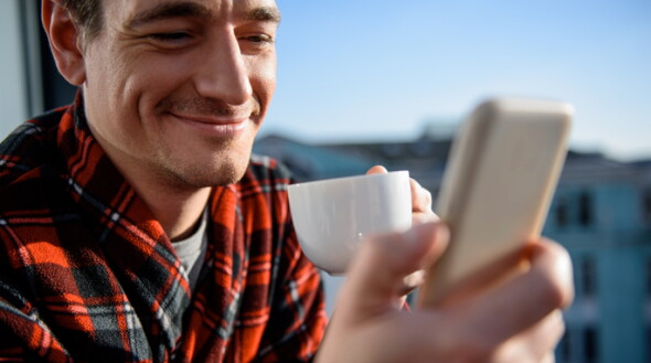 man wearing red and black checkered shirt smiling at his mobile holding a cup in his hand