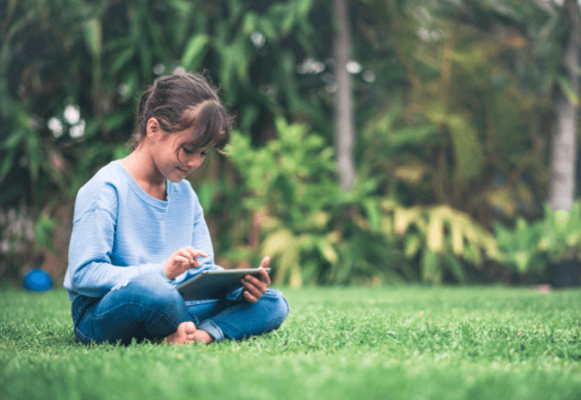 Young girl playing on a tablet in a grassy outdoor area.