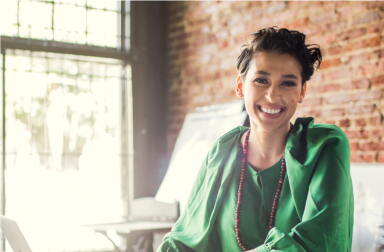 A smiling woman sitting inside a sun-lit office.